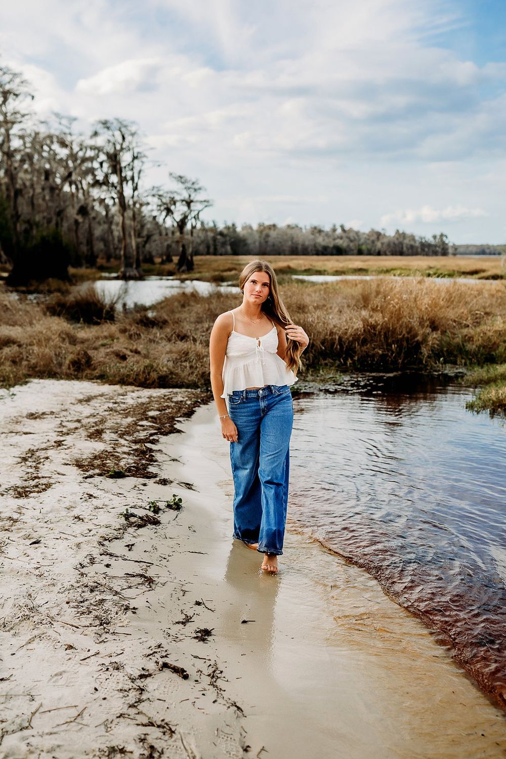 Young woman in white top and flared jeans walking barefoot along a sandy marsh shoreline under a cloudy sky.