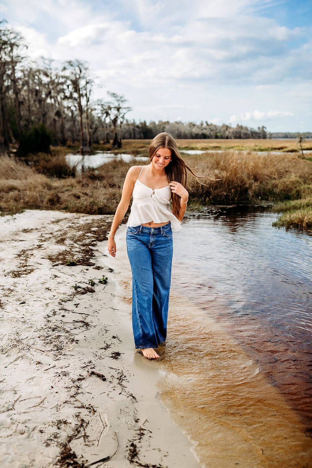 Young woman in white top and flared jeans walking barefoot along a shallow sandy creek in a natural wetland.
