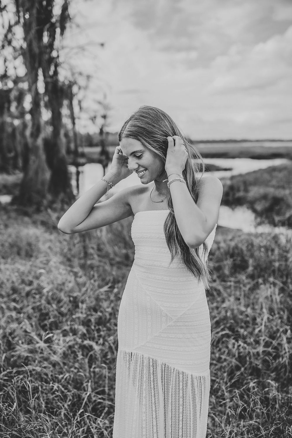 Black and white photo of a smiling young woman in a white maxi dress posing outdoors near a marsh.