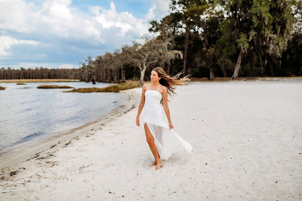 A woman in a flowing white dress walks barefoot on a sandy beach with trees and water in the background.