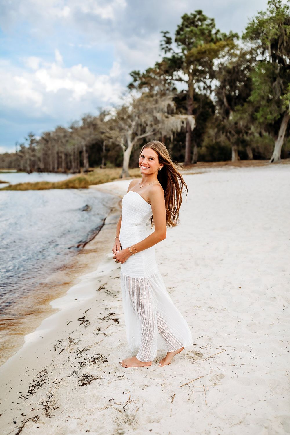 Young woman in white dress walking barefoot on sandy beach shore with trees in background.