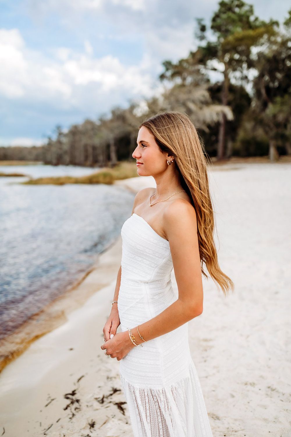 Young woman in white strapless dress standing on sandy beach shore with trees and cloudy sky backdrop.