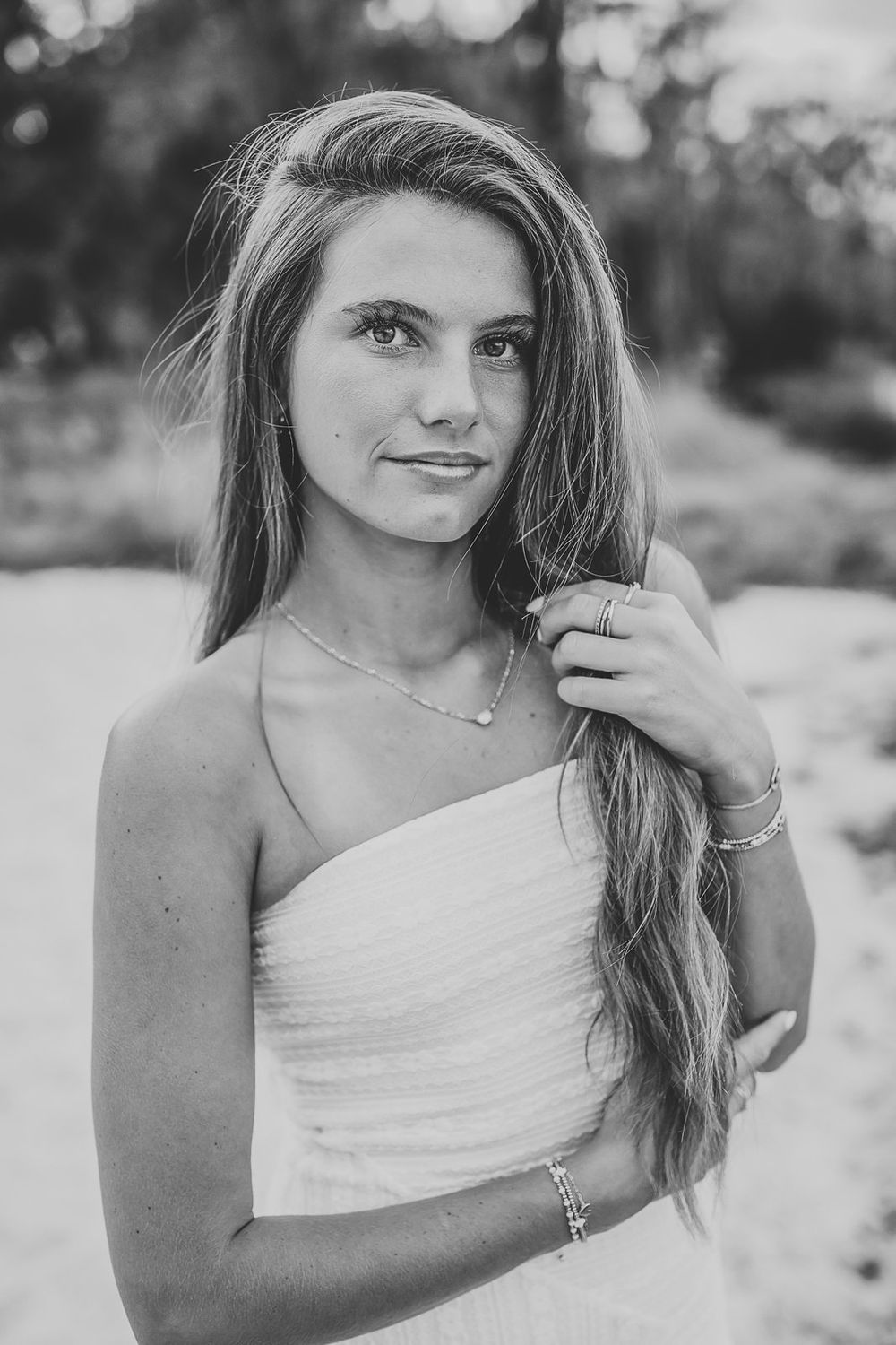 Black and white portrait of a young woman in a white strapless dress, holding her long hair, wearing delicate jewelry.