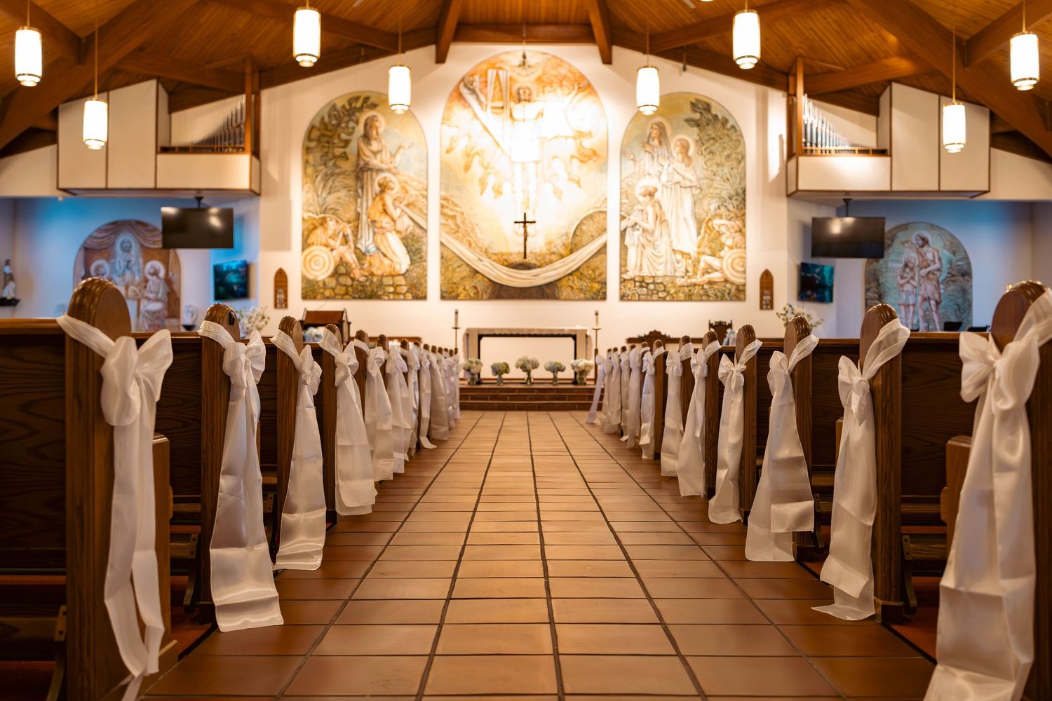 Elegant church aisle decorated with white satin bows on wooden pews, leading to a beautiful altar with religious murals.