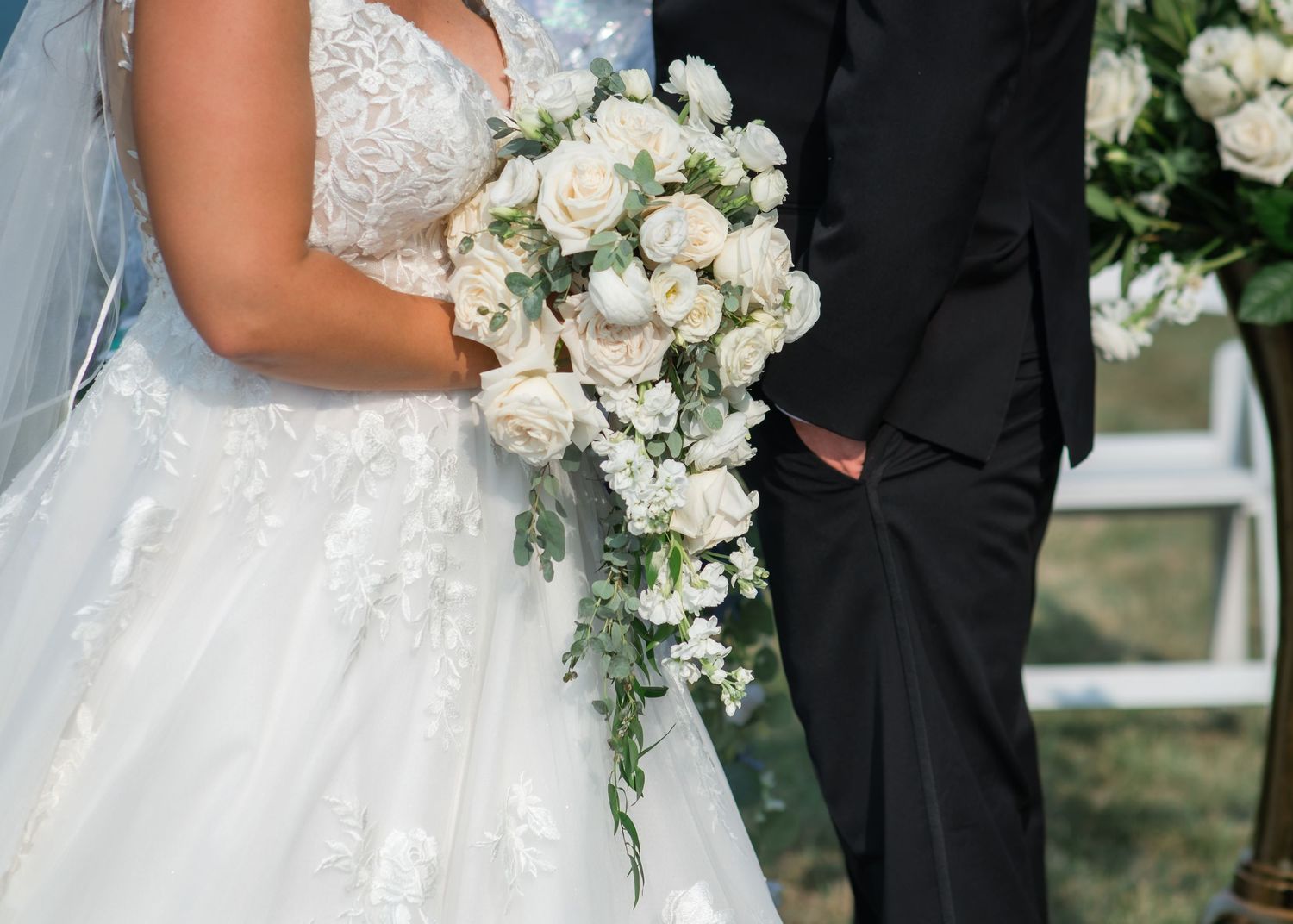 Bride in lace wedding gown holding cascading white rose bouquet standing beside groom in black suit.