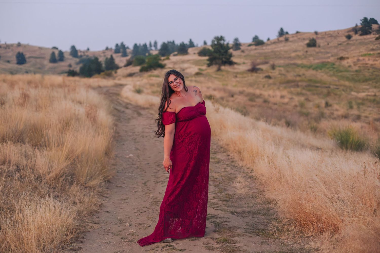 Pregnant woman in red off-shoulder gown poses on a dirt path surrounded by golden hills.