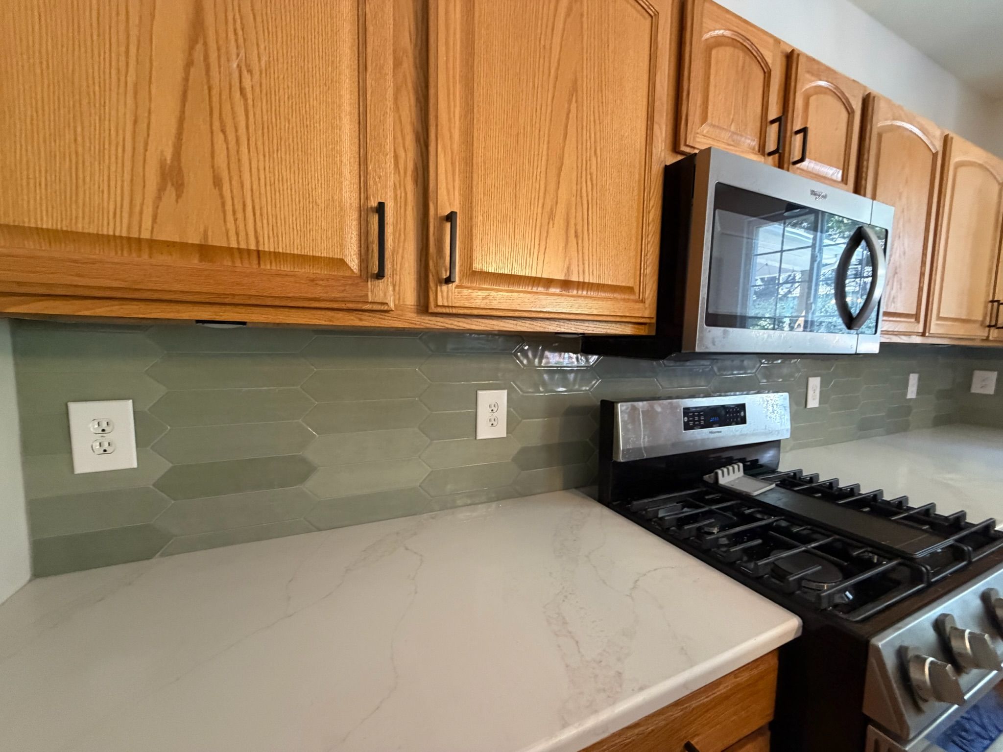 Oak kitchen cabinets with sage green elongated hexagon glass tile backsplash and white quartz countertop near gas range.