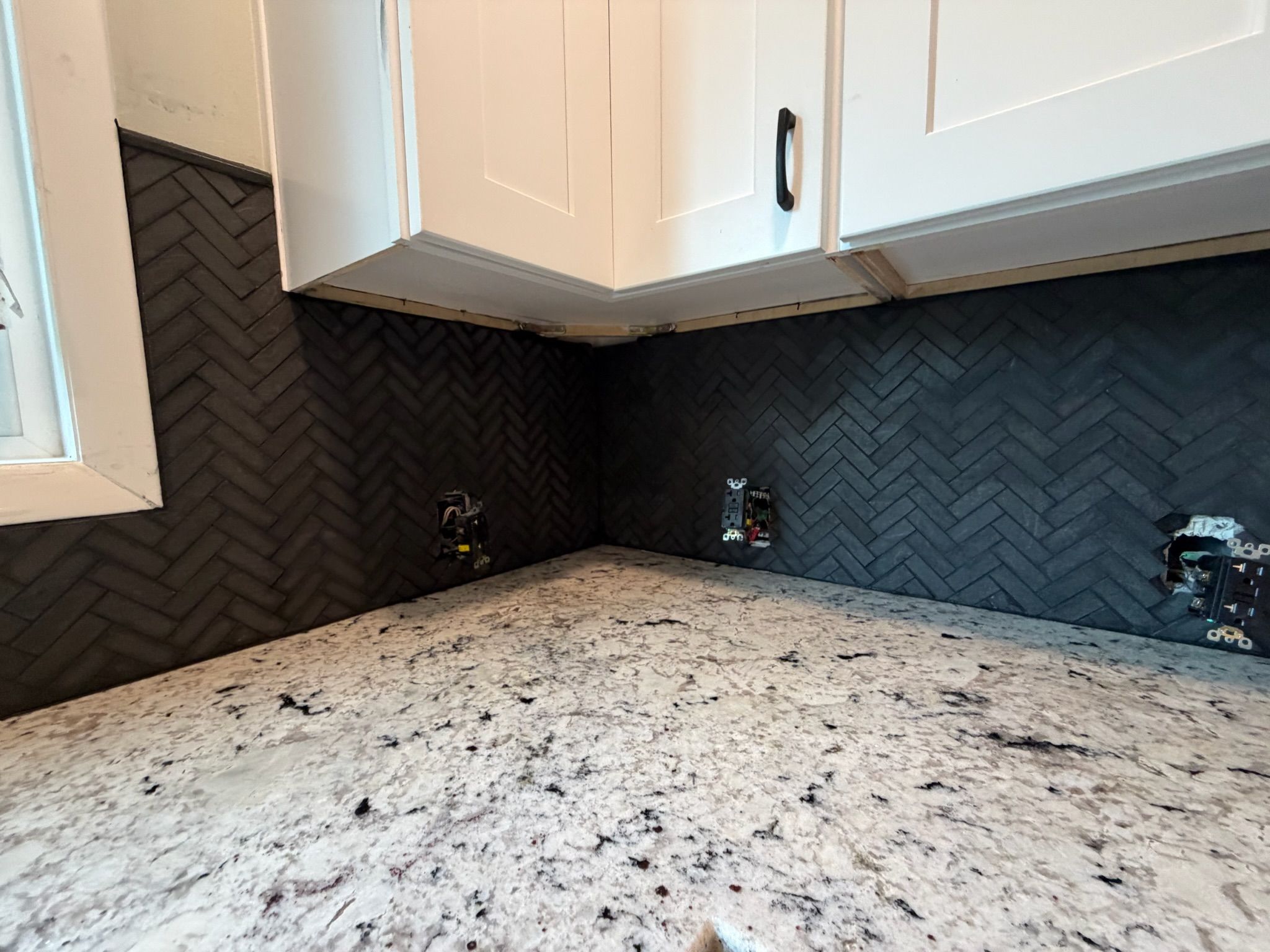 Dark herringbone tile backsplash with white shaker cabinets and granite countertop in modern kitchen.