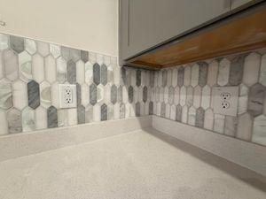 Close-up of elongated hexagon marble mosaic backsplash tiles in gray and white tones beneath gray kitchen cabinets.