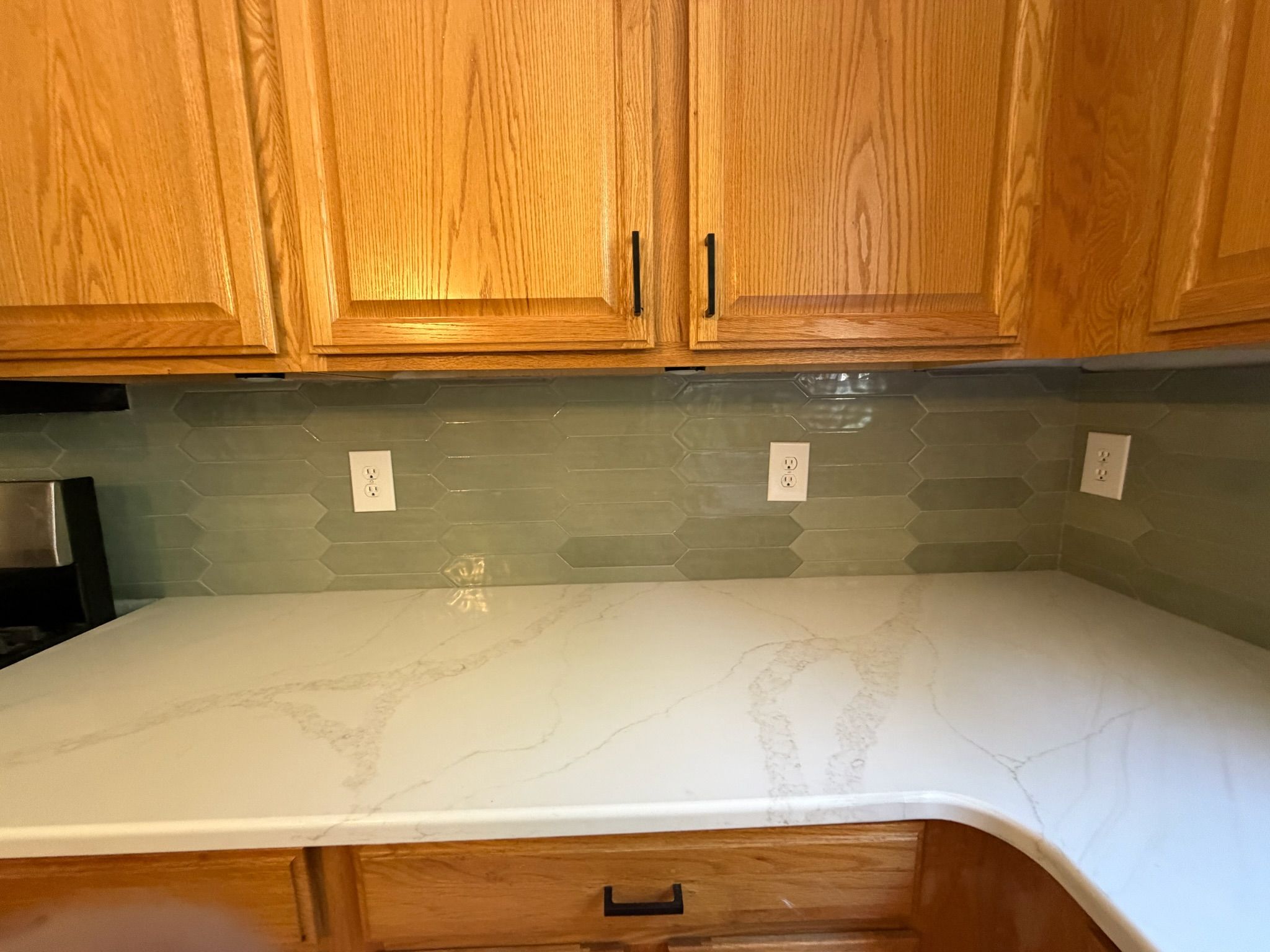 White marble quartz countertop with sage green glass tile backsplash and oak kitchen cabinets with black hardware.