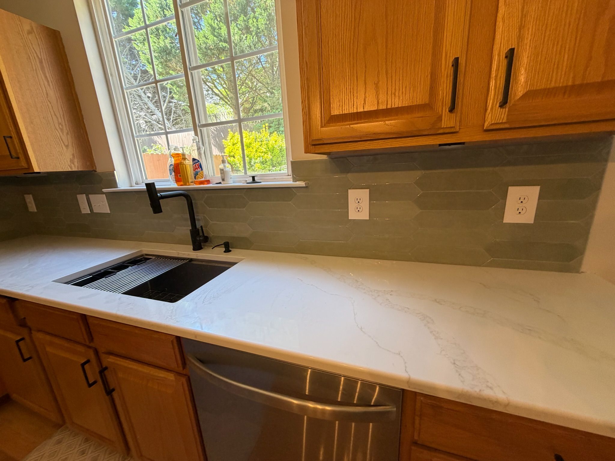 Kitchen remodel with warm oak cabinets, black farmhouse sink, sage green glass tile backsplash, and white quartz countertop.