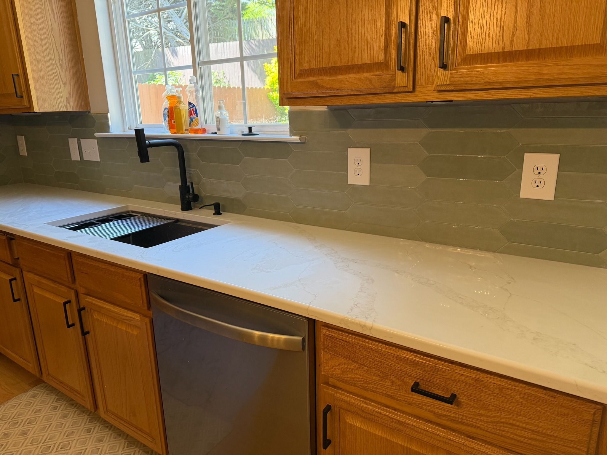 White marble quartz countertop with green subway tile backsplash, black farmhouse sink, and stainless dishwasher.