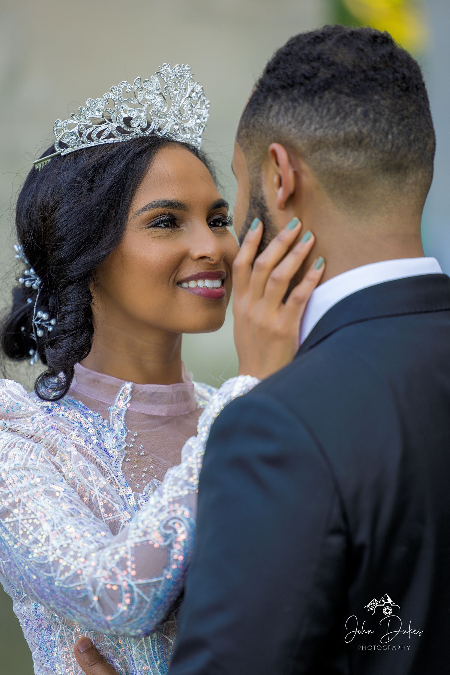 A bride wearing a sparkling tiara and beaded gown smiles at a groom in a black suit during a romantic wedding moment.