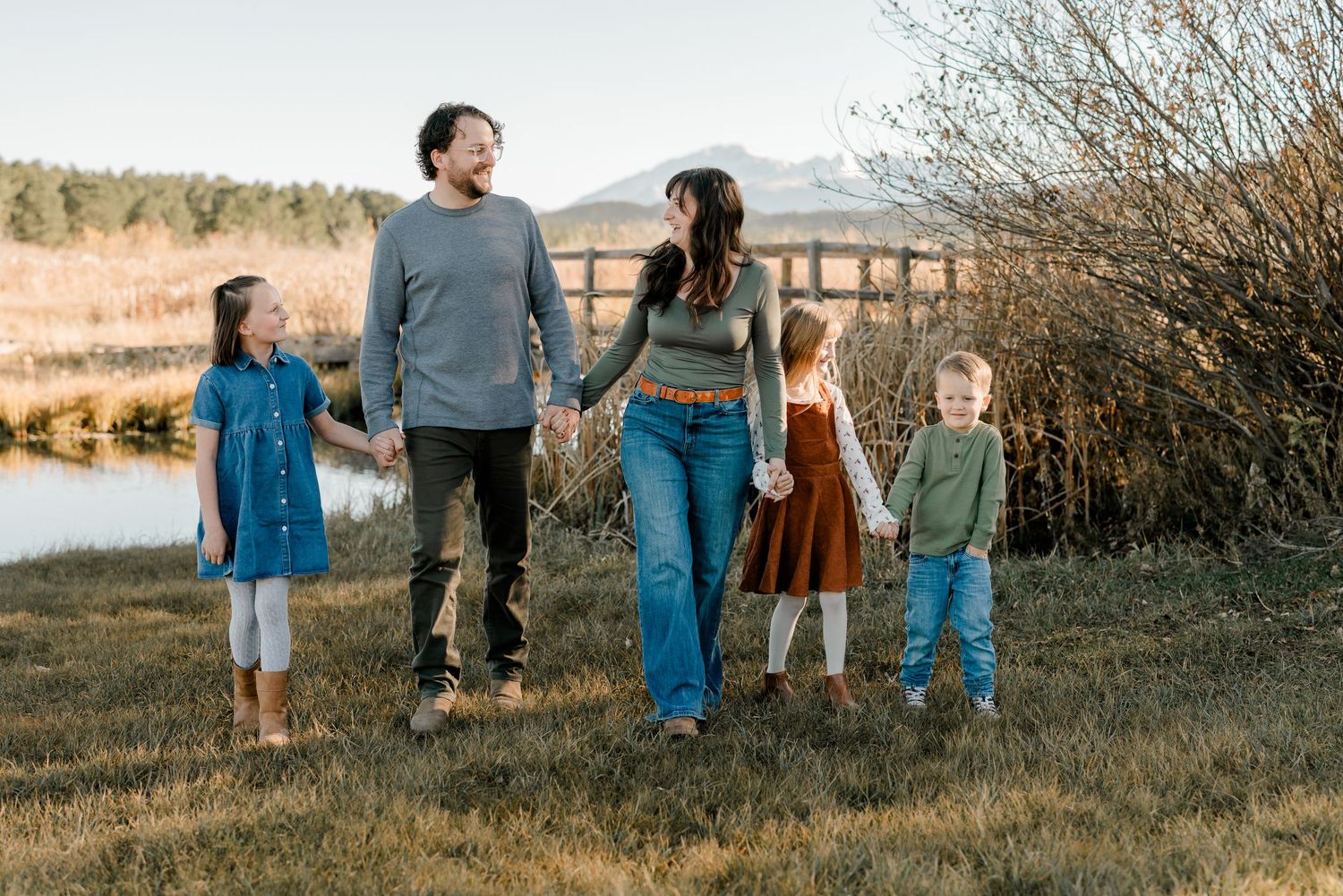 Happy family of five holding hands while walking through a scenic meadow near a pond on a sunny autumn day.