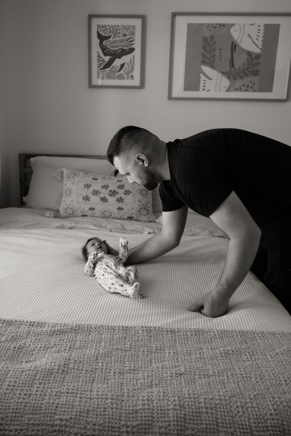 Black and white photo of a father gently placing his newborn baby on a bed in a cozy bedroom.