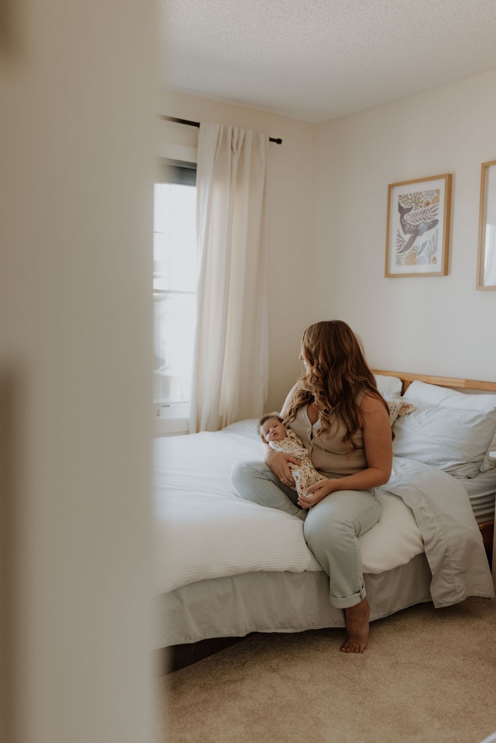 A mother sits on a bed cradling her newborn baby in a softly lit bedroom with white curtains and neutral decor.