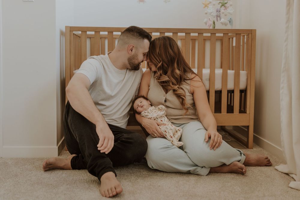 A couple sits on nursery floor with newborn baby in front of wooden crib while their photographer captures a special moment between them