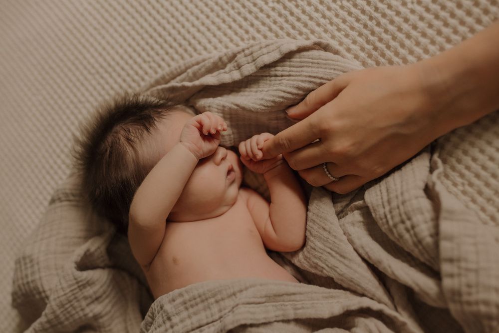 Newborn baby sleeping on a soft beige muslin blanket while an adult gently touches its tiny hand.