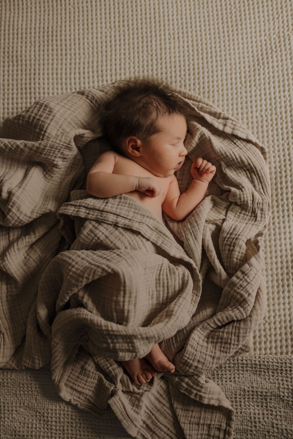Peaceful newborn baby sleeping on soft beige muslin blanket, captured in warm natural light photography.