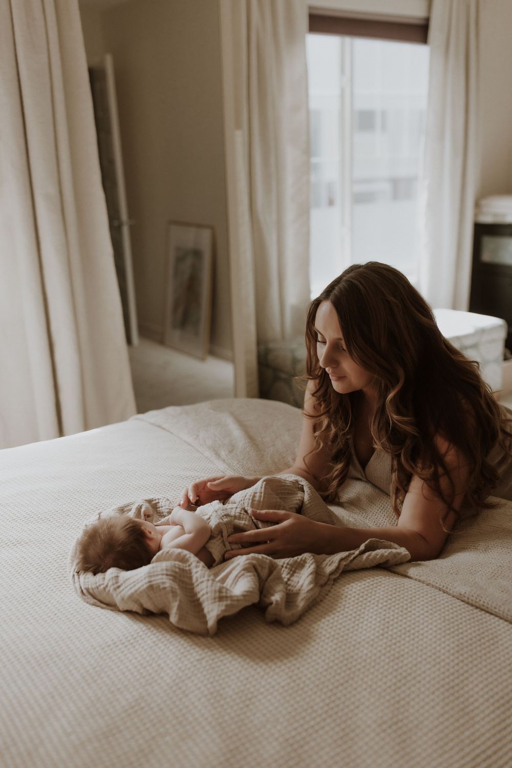 Mother lying on white bed lovingly gazing at her newborn baby wrapped in a cozy blanket.