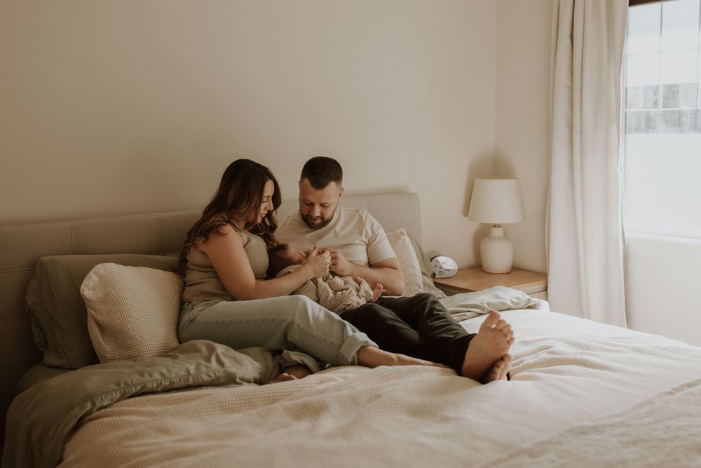 Couple sitting on a bed together, smiling and looking at a newborn baby in a cozy bedroom in calgary during thier in home newborn photoshoot