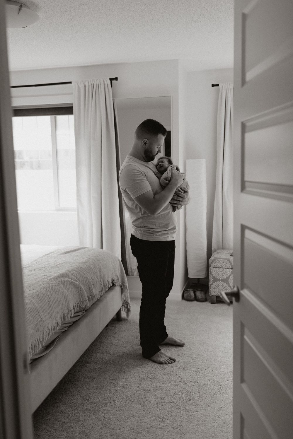 Black and white photo of a father holding his newborn baby in his bedroom, as a calgary photographer captures a special bonded moment