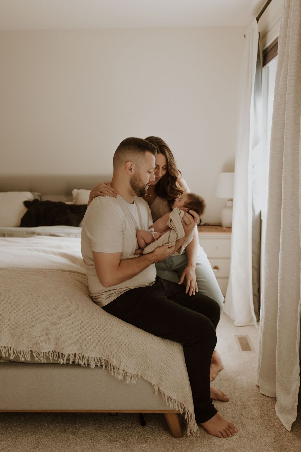 A couple sits on a bed, lovingly gazing at their newborn baby in a bright, cozy bedroom in calgary alberta during their newborn photoshoot