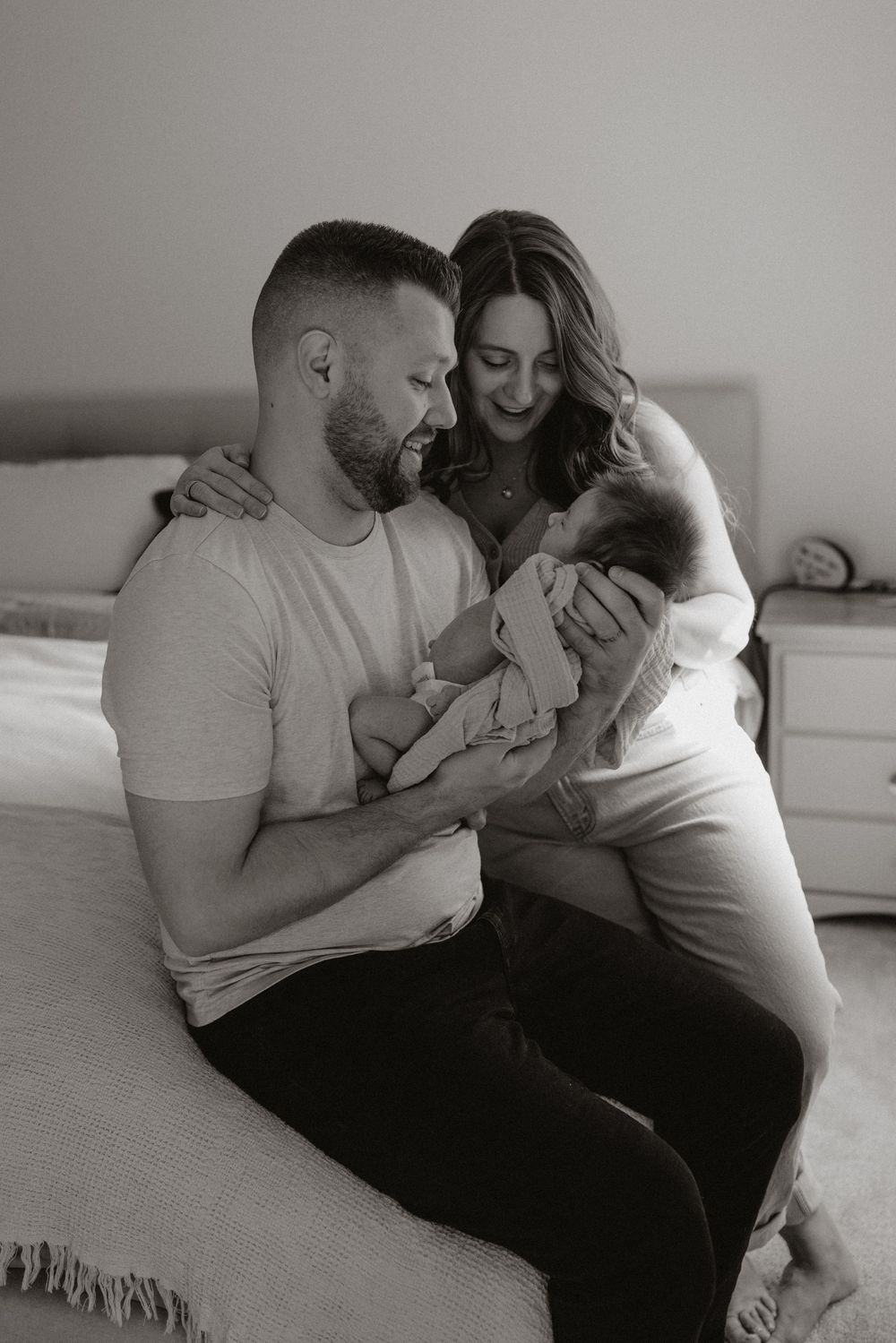 Black and white photo of parents sitting on bed lovingly gazing at their newborn baby together.