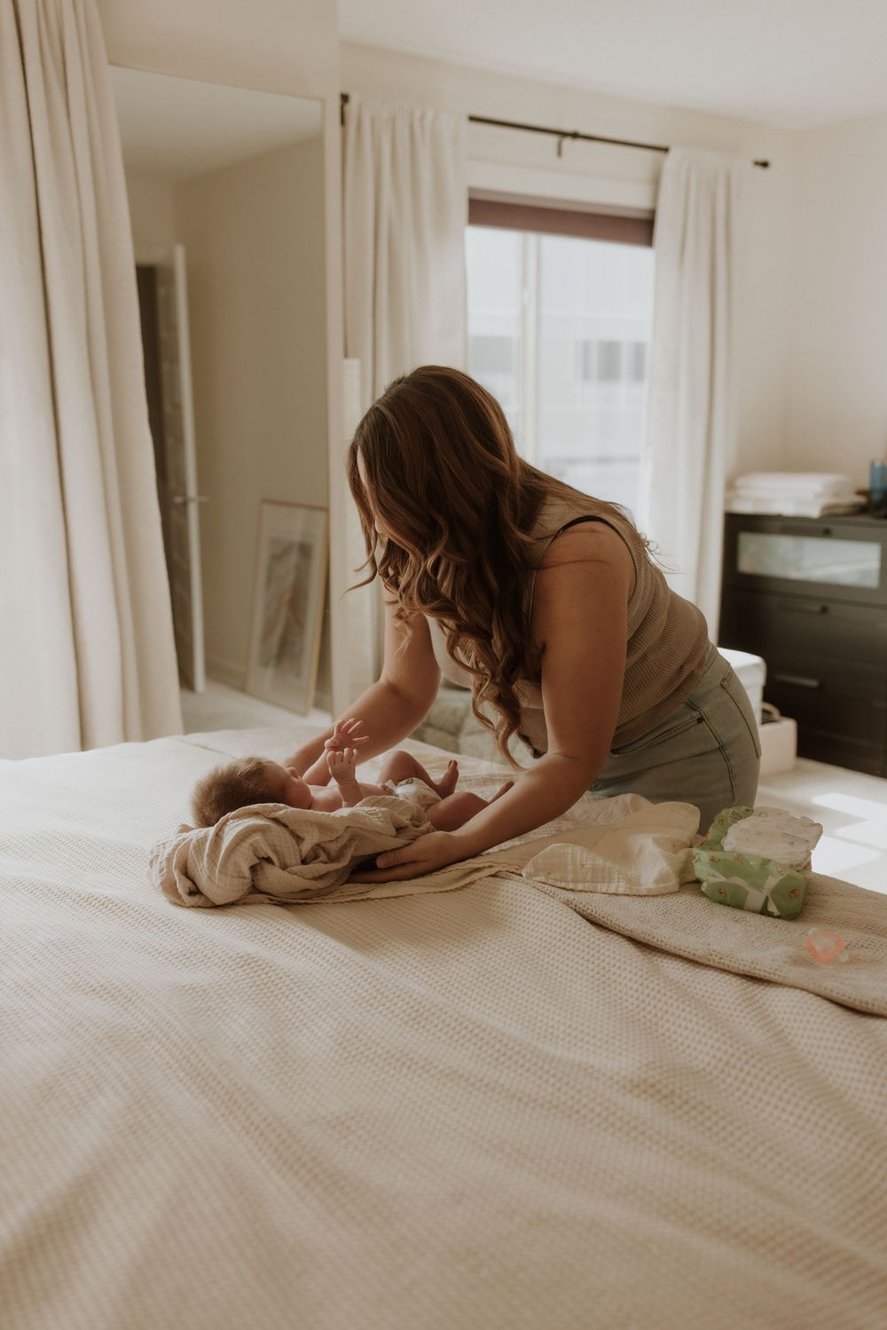 A mother tenderly cares for her newborn baby on a white bed in a bright, softly lit bedroom.