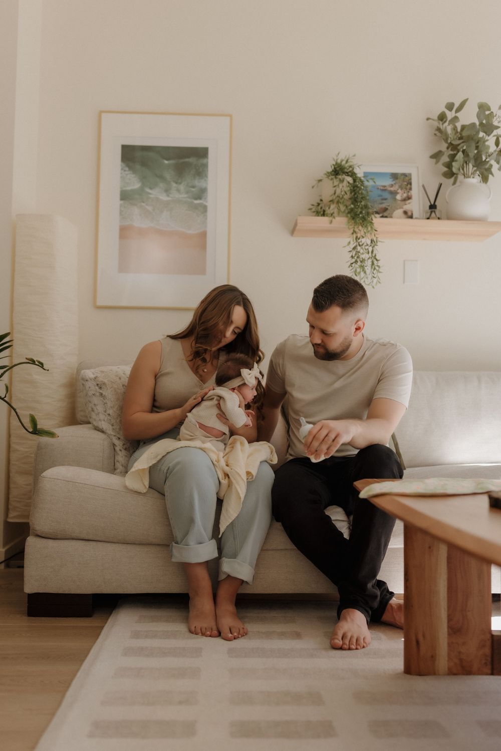 A young family sits together on a cream sofa, with a mother holding her newborn baby while the father looks on lovingly.