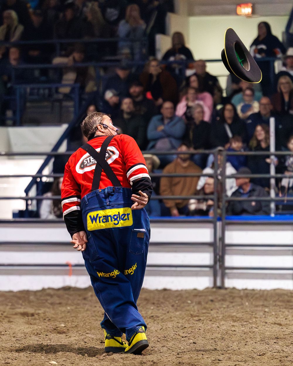 Rodeo clown in Wrangler overalls tosses hat in the air before a crowd.