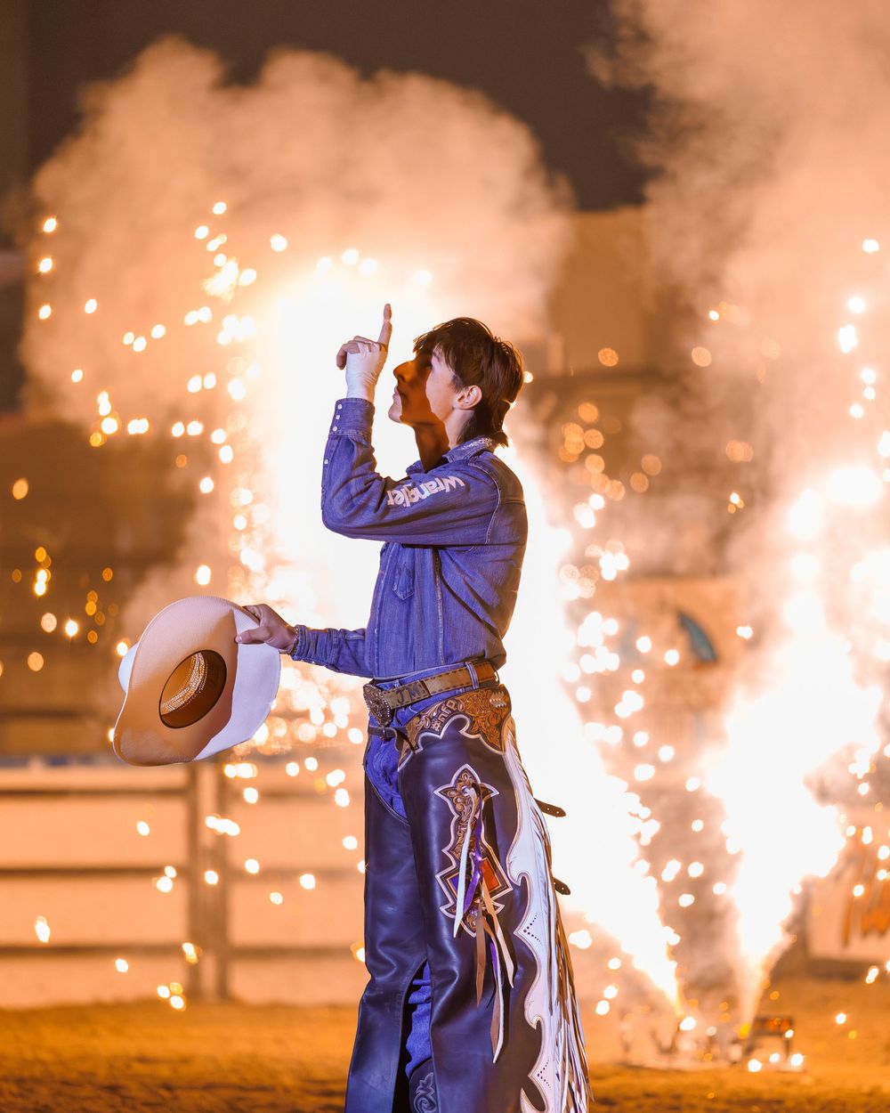 Cowboy in blue shirt raises finger triumphantly amid fireworks at a rodeo event.