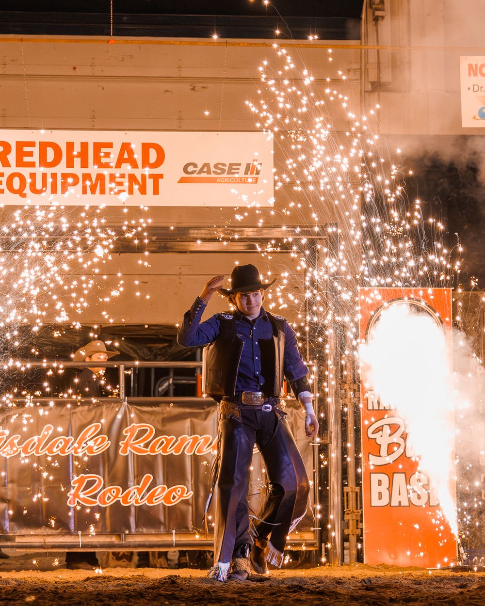 Cowboy stands dramatically in front of pyrotechnic fireworks at Redhead Equipment rodeo.