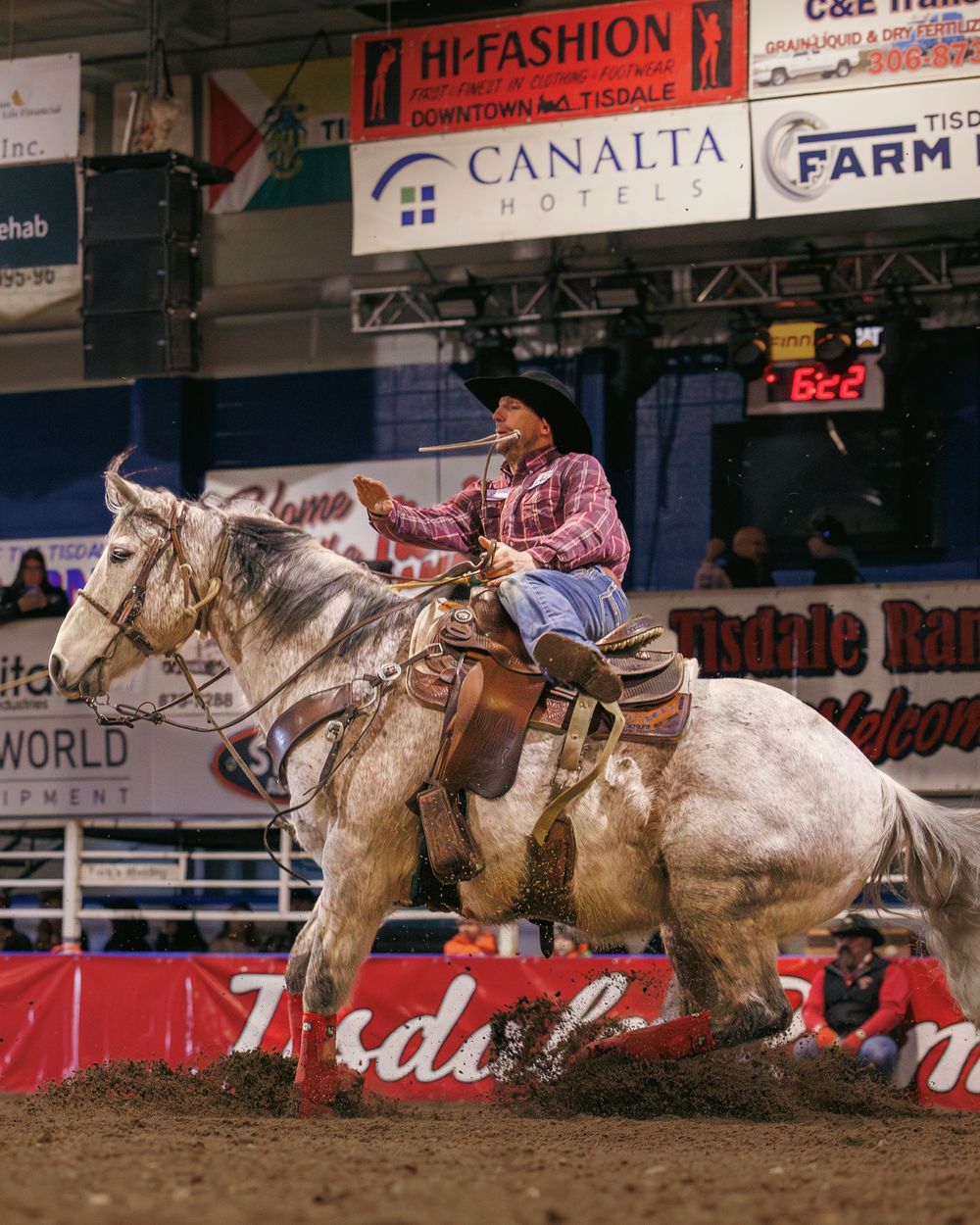 Female cowboy on grey horse competes in fast-paced indoor rodeo barrel racing event.