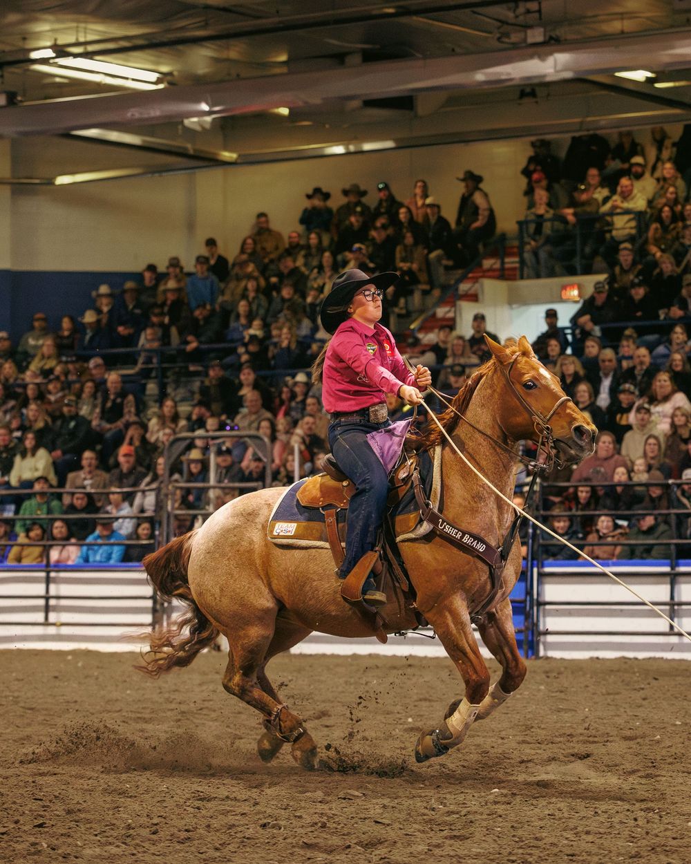 Cowgirl in pink shirt rides palomino horse at full speed during indoor rodeo event.