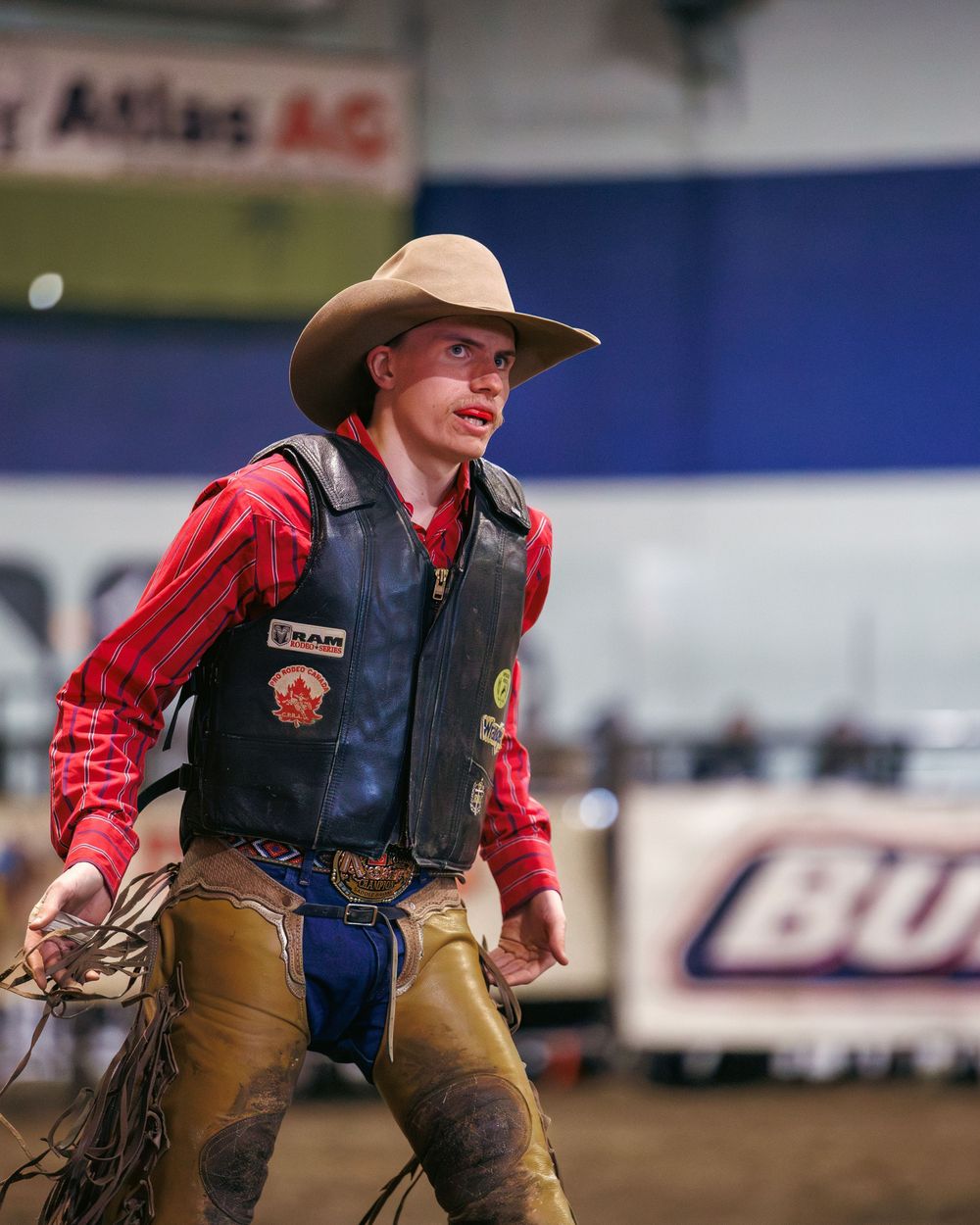 Young rodeo cowboy in leather vest and red shirt walks arena after competition event.