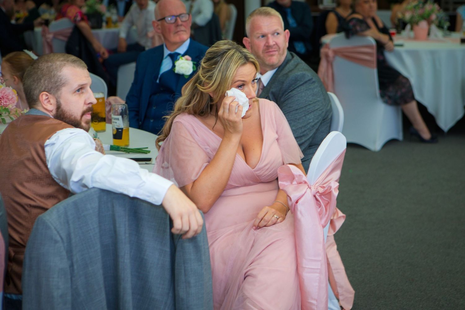 Woman in pink dress crying at wedding reception, dabbing eyes with tissue while seated at decorated table.