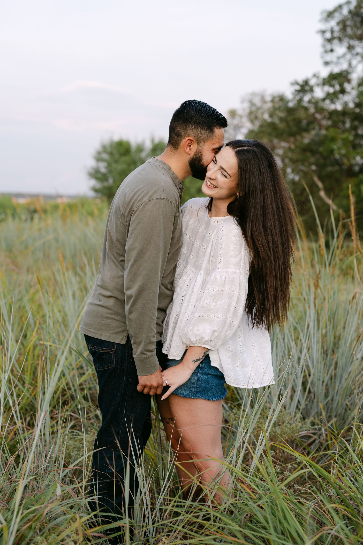A couple shares a romantic moment in a lush green field, the man kissing her forehead as she smiles softly.