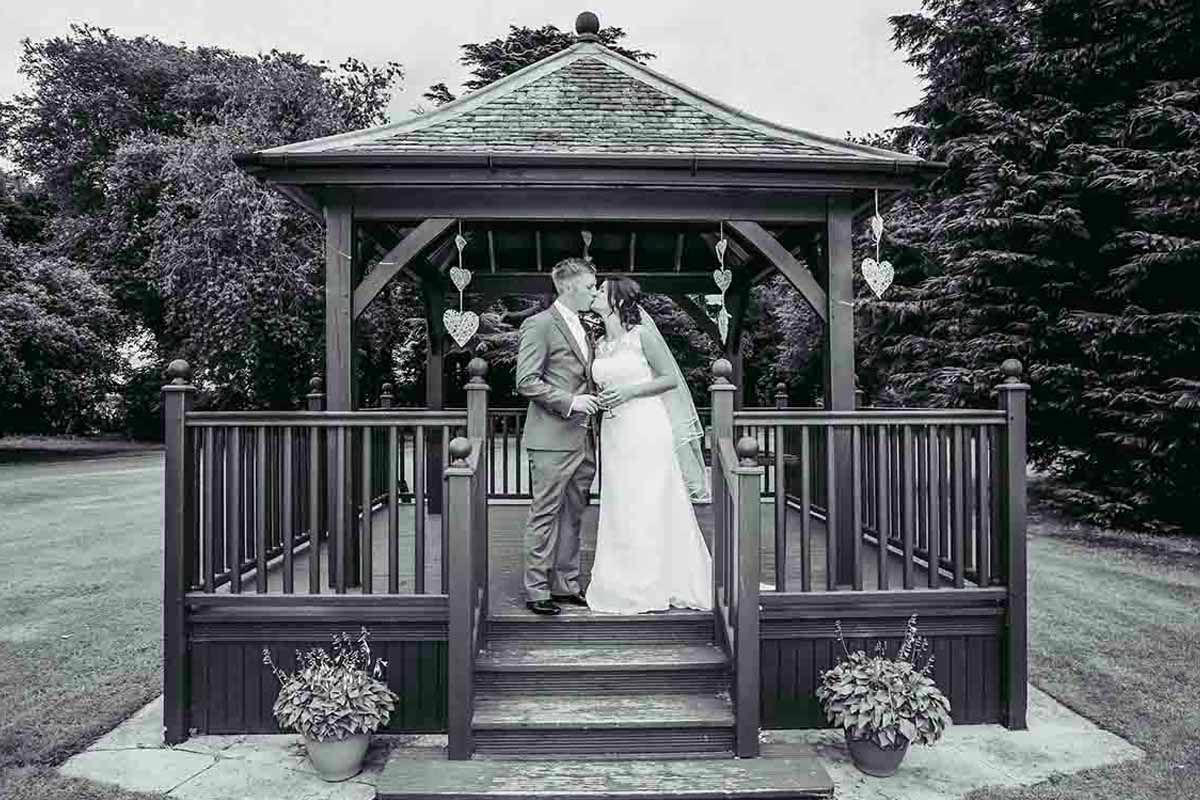 A bride and groom share a romantic kiss on the steps of an elegant wooden gazebo at Greenhill Hotel - Wigton- Cumbria