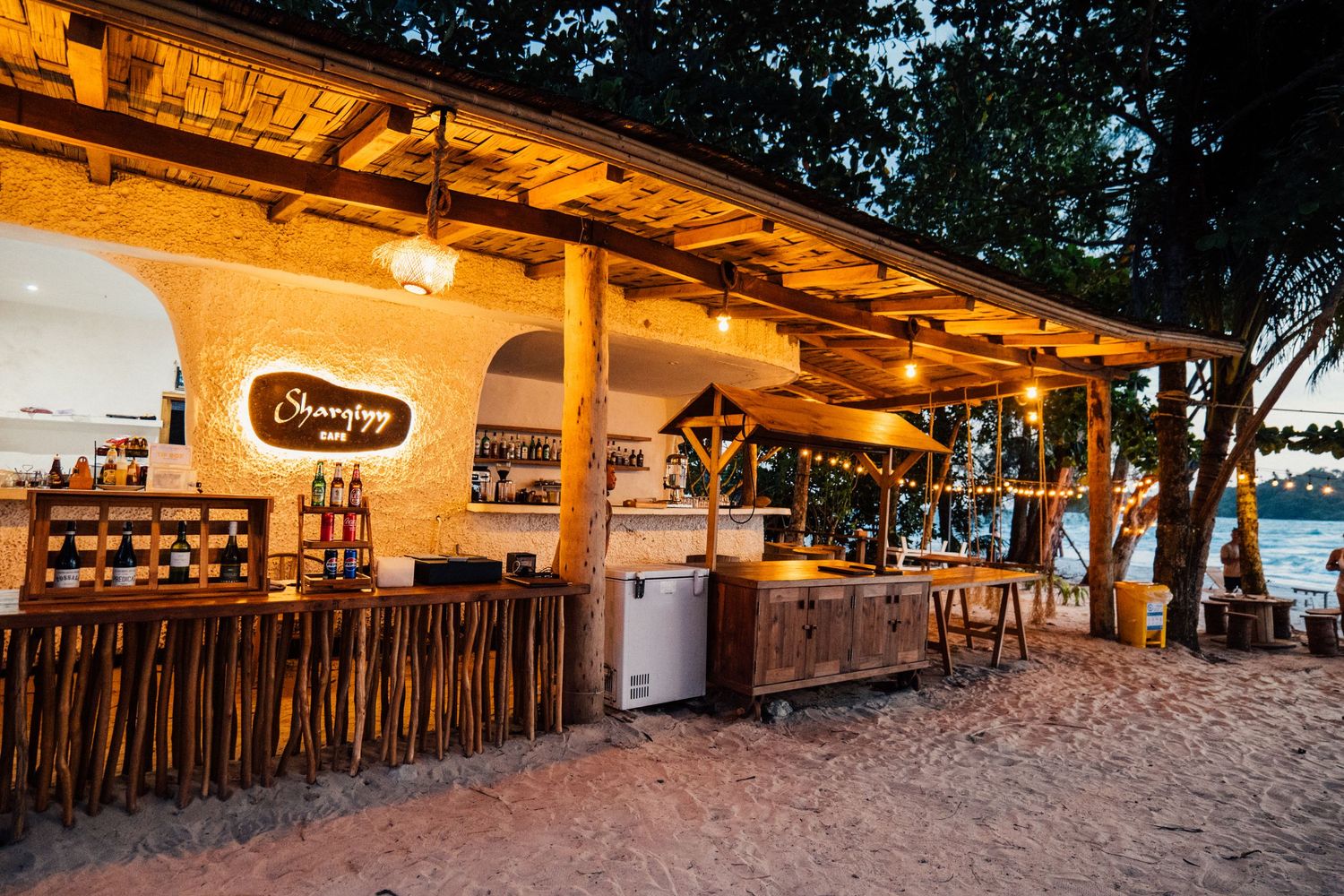Cozy beachside bar with warm lighting, bamboo decor, and sandy floors at twilight.