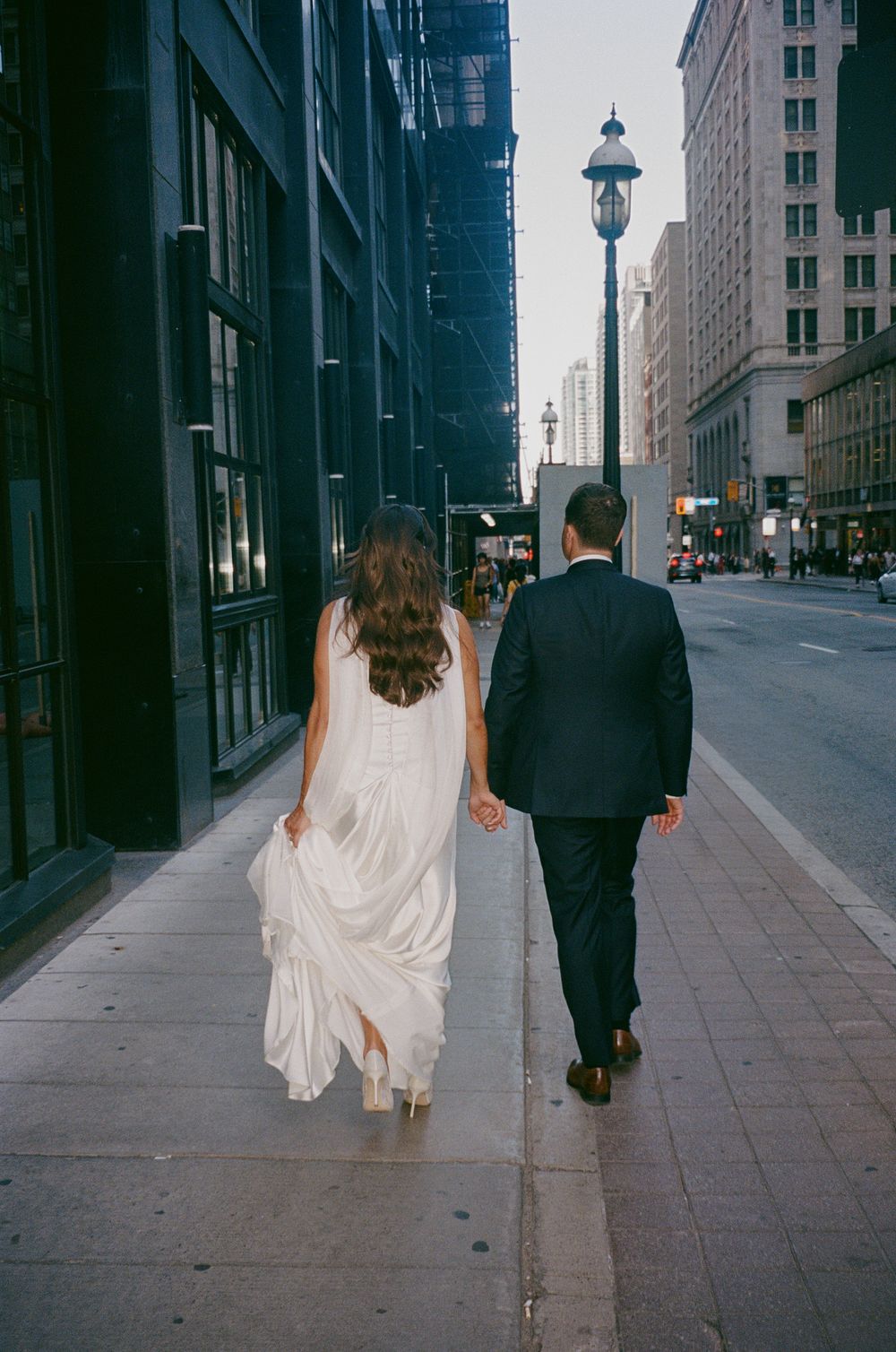 Bride and groom walk hand in hand down a city sidewalk, bride's white gown flowing behind her.