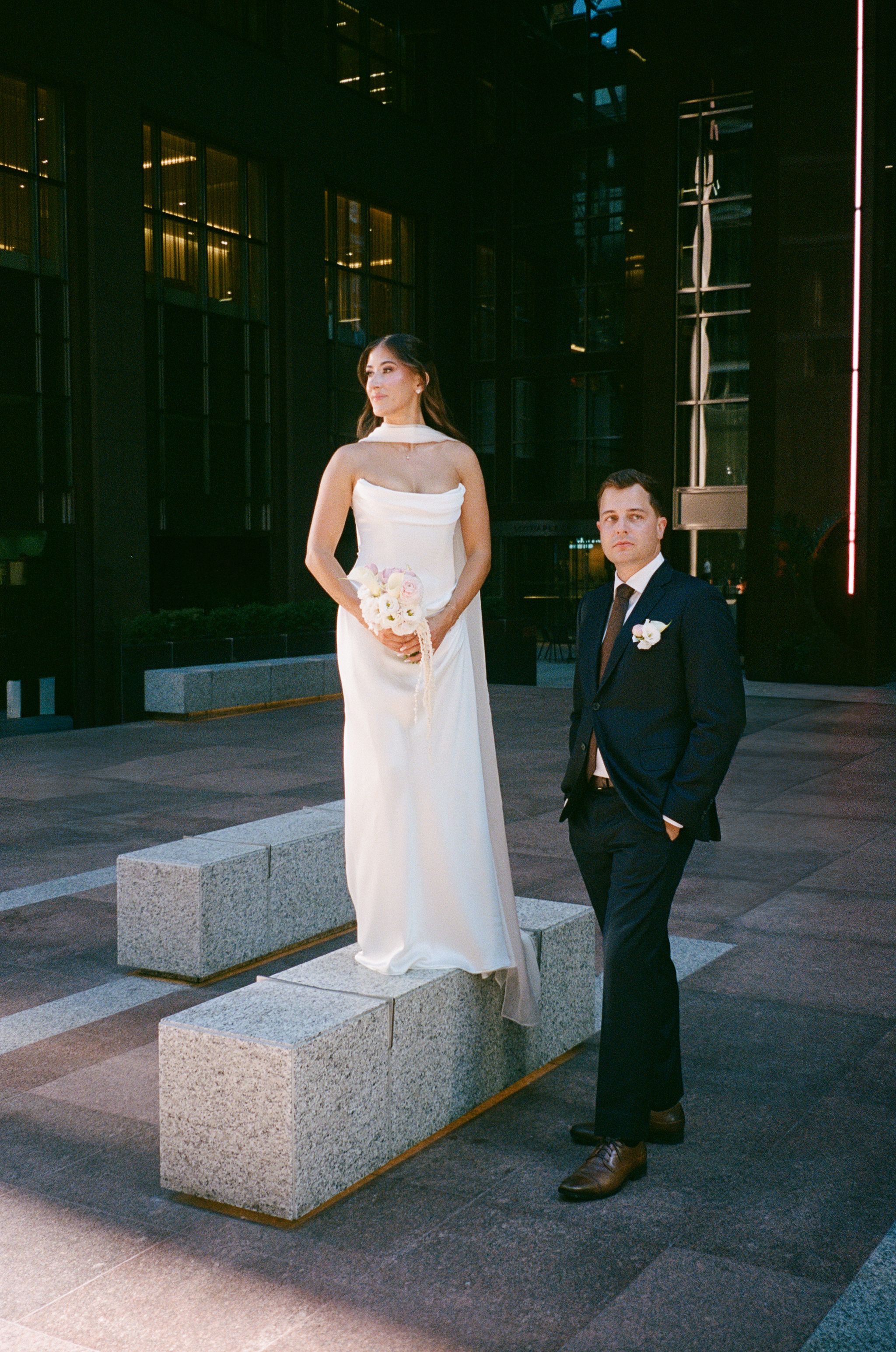Bride stands elevated on concrete blocks beside groom in dramatic evening light outside modern building.