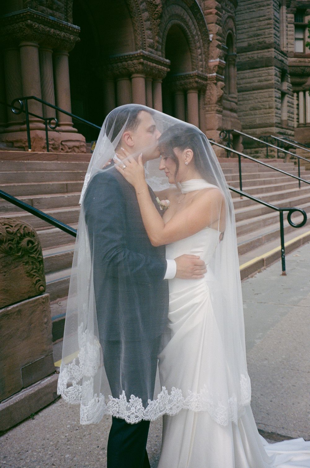 Bride and groom share a romantic kiss beneath a lace-trimmed veil on church steps after ceremony.