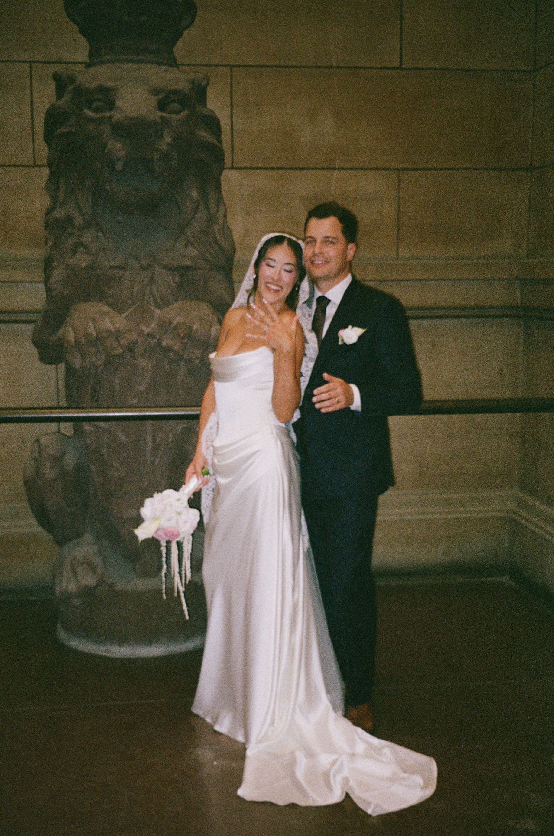 Bride and groom pose elegantly before a stone lion sculpture inside a grand historic building.