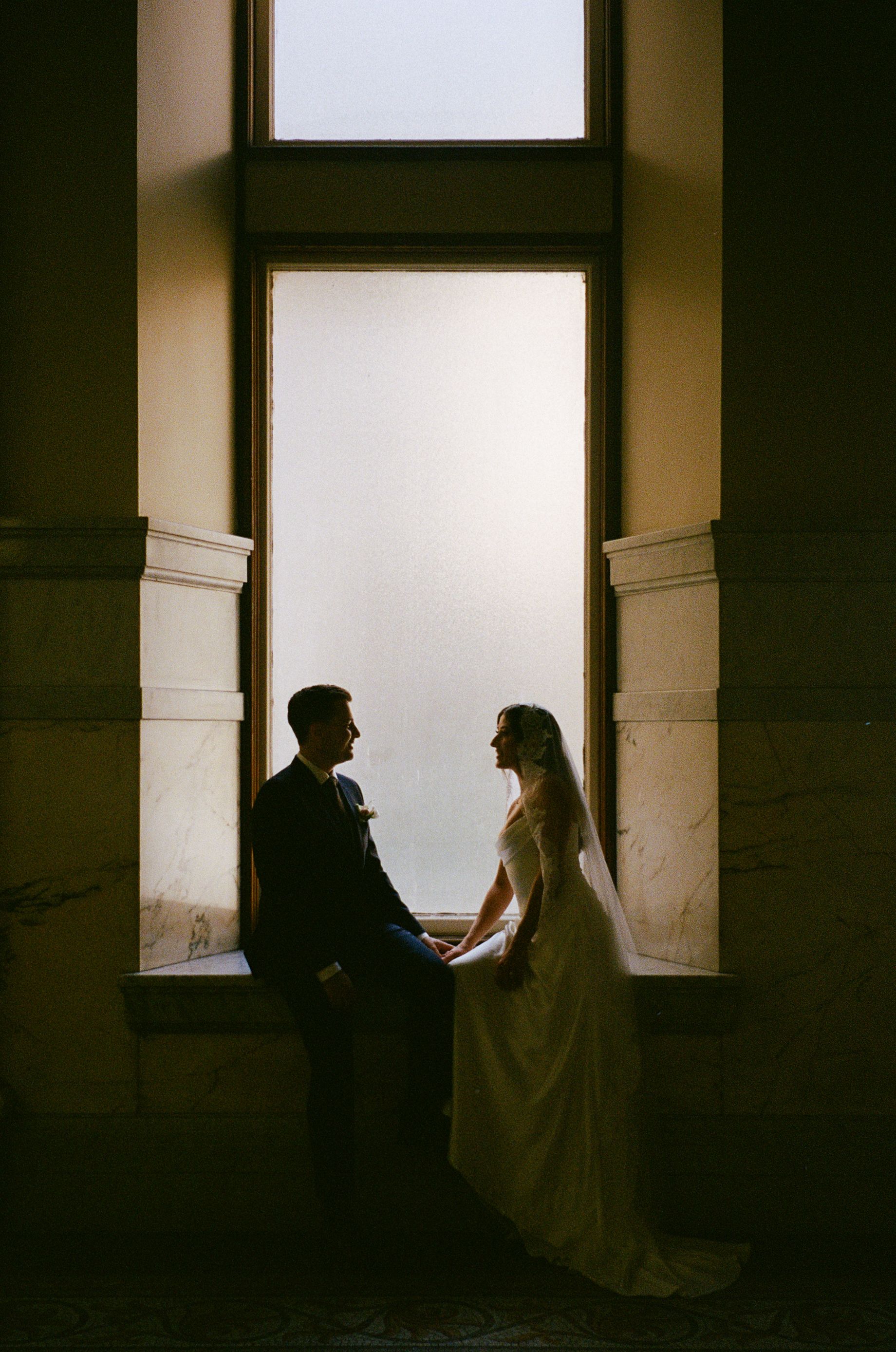 Silhouette of bride and groom holding hands by a tall arched window in a historic venue.