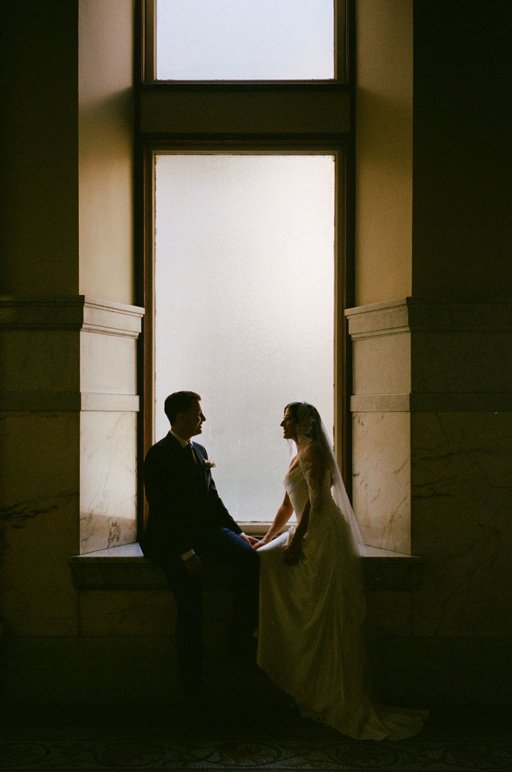 Silhouette of bride and groom holding hands by a tall arched window in a historic venue.