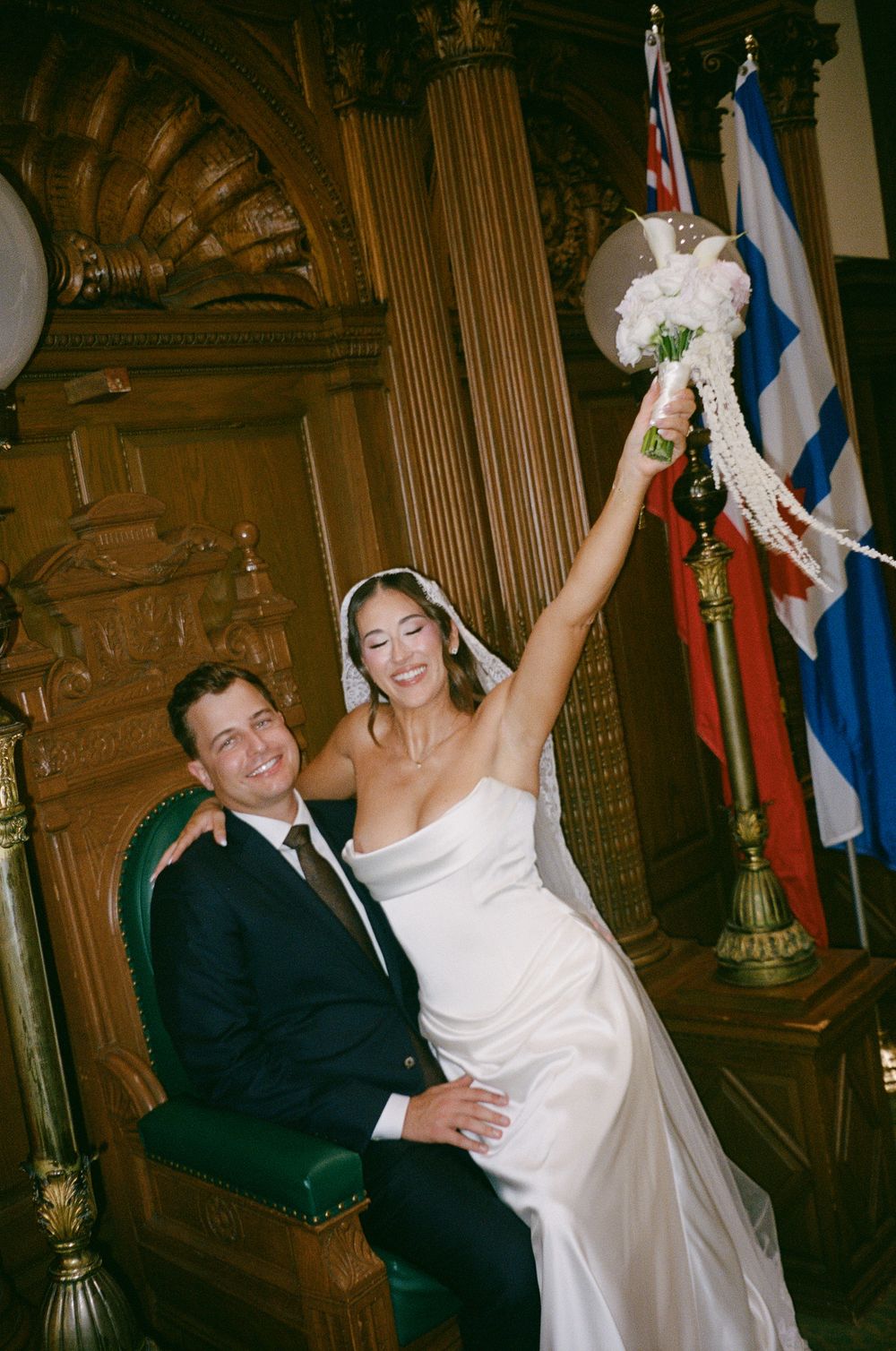 Joyful bride raises bouquet while groom sits in ornate wooden chair during wedding ceremony with flags in background.