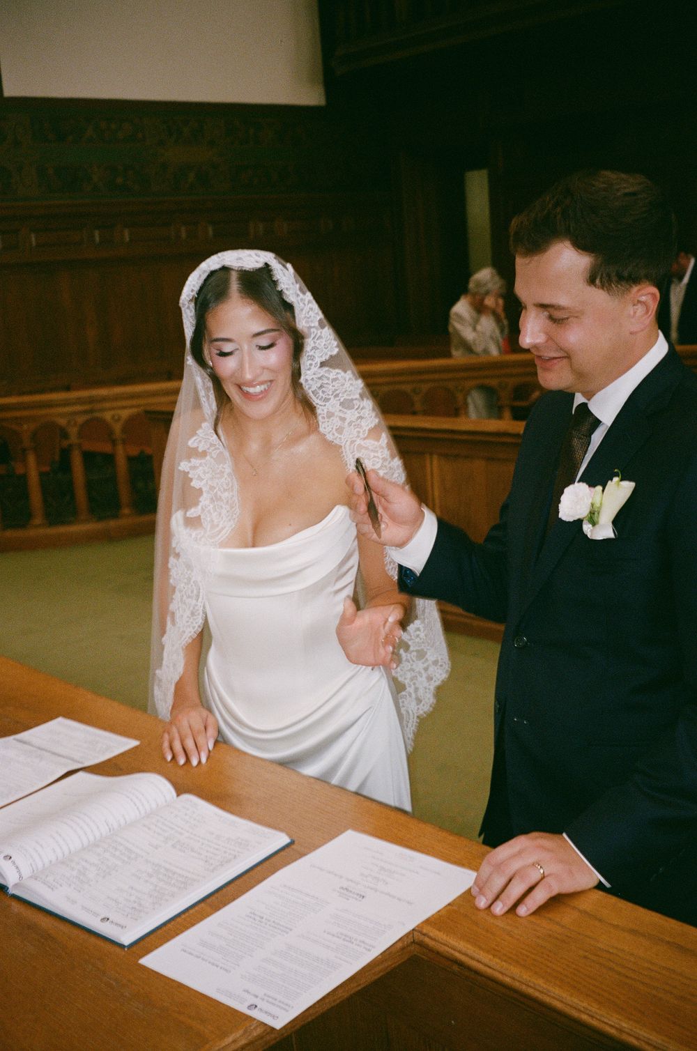 Smiling bride and groom sign wedding registry documents at wooden table during civil ceremony.