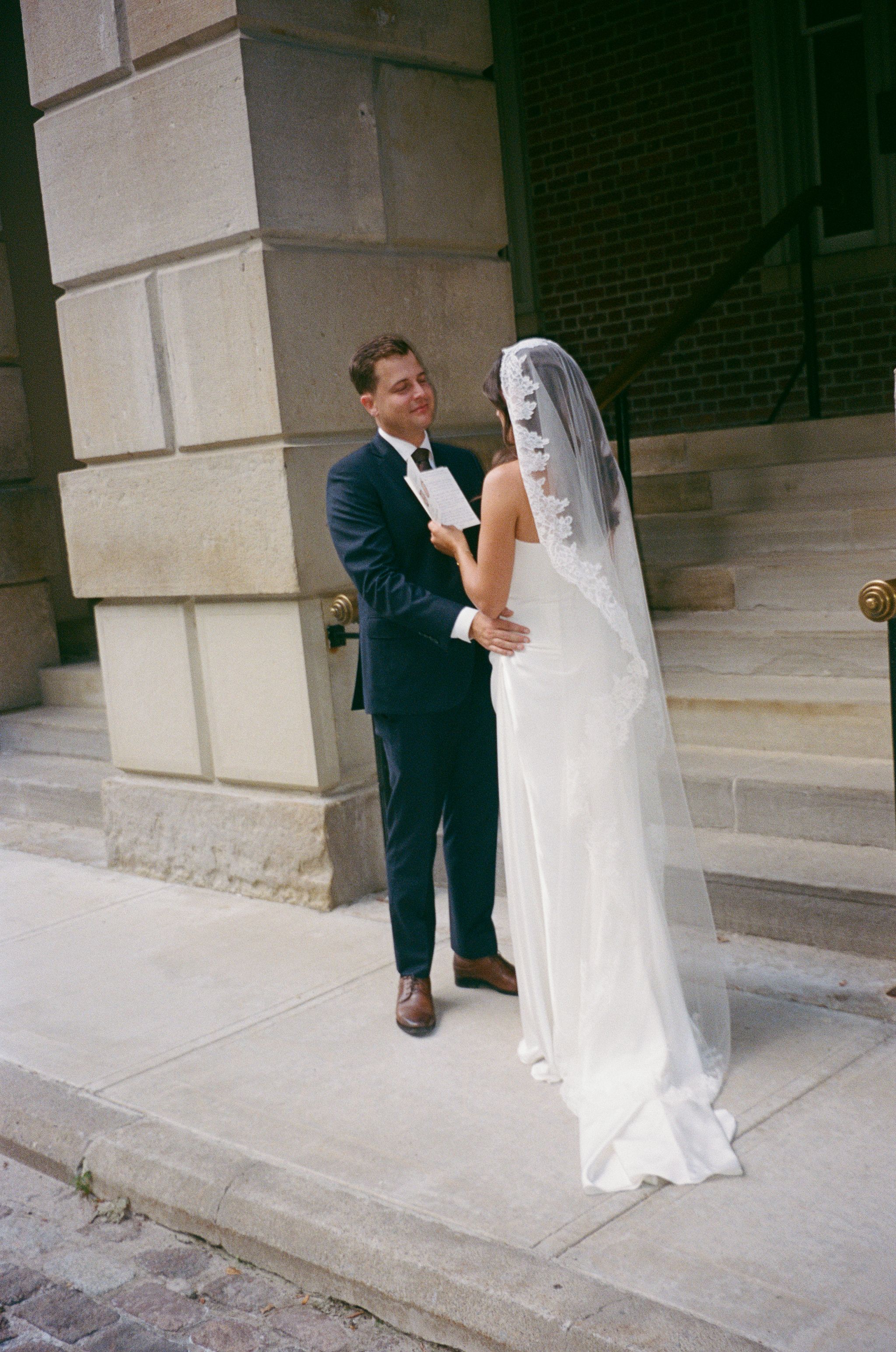 Groom reads vows to bride on stone steps outside historic building during intimate wedding ceremony.