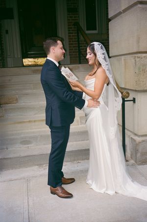 Bride and groom share vows on stone steps outside classic building, groom holding ceremony booklet.